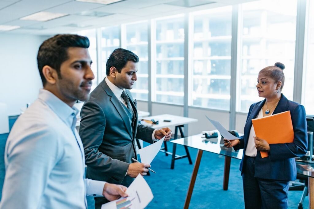 Three colleagues in business dress stand with printed documents in a glass-walled meeting room overlooking the city.