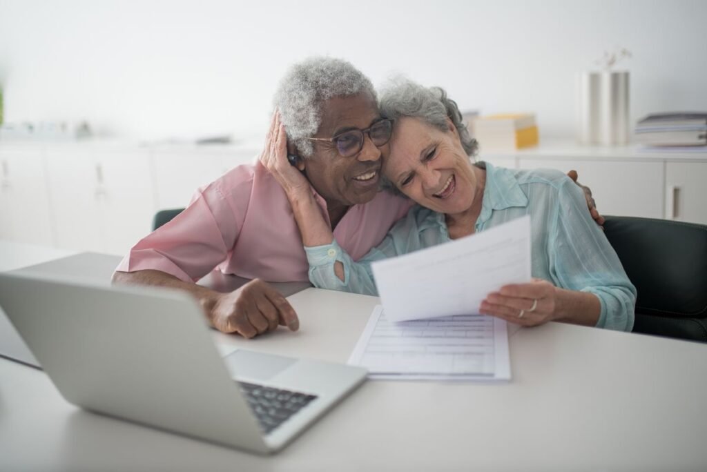 An older couple sit close together at a table with a laptop, smiling as they read through a printed statement.